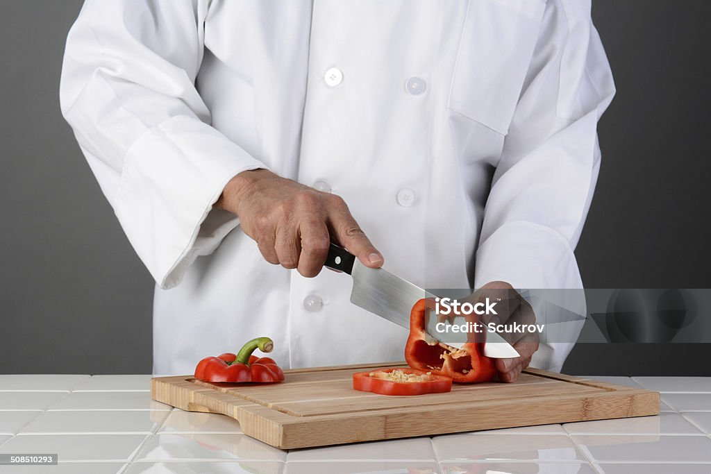 Chef Slicing Red Bell Pepper Closeup of a chef slicing a red bell pepper on a wood cutting board. Horizontal format on a light to dark gray background. Man is unrecognizable. Model Released. Adult Stock Photo