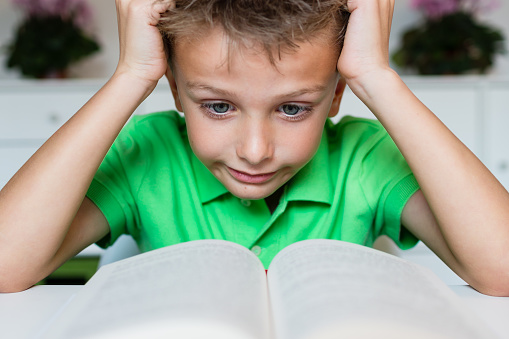Young boy in green polo shirt having serious learning difficulties while trying to read a textbook from school.