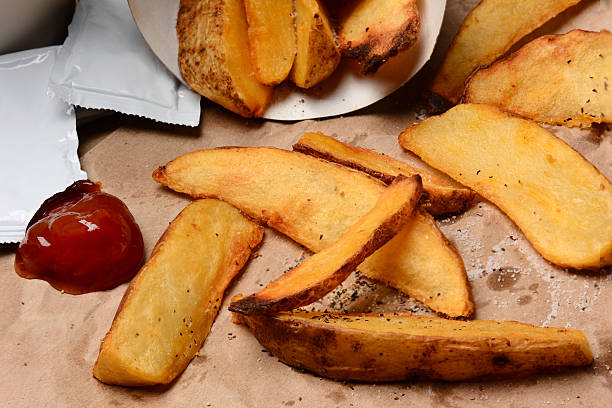 Closeup of  some French Fries spilled onto a brown bag. Ketchup dollop and packets  with salt and pepper. Horizontal format filling the frame.