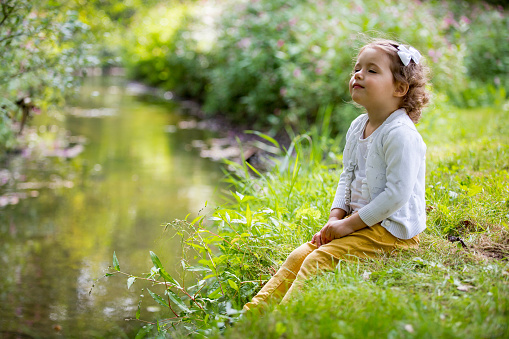 Sweet, happy little girl sitting on a grass in a park at a spring stream with flower in hand. Laughing, enjoying fresh air in forrest.