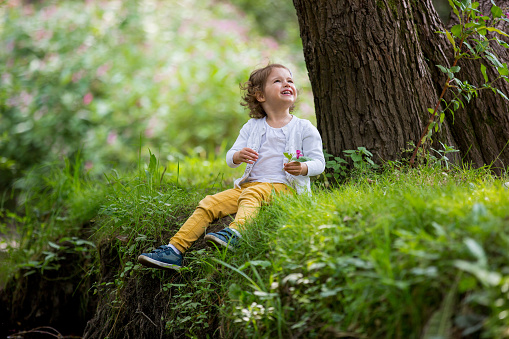 Sweet, happy little girl sitting on a grass in a park at a spring stream with flower in hand. Laughing, enjoying fresh air in forrest.