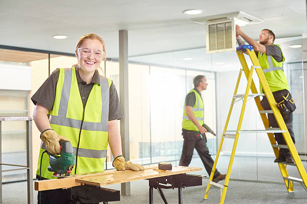 female apprentice carpenter on site a teenage female carpenter is sawing studs on her workbench in a modern office refurbishment. IN the background a co-worker can be seen up a ladder working on the air conditioning unit. They are wearing safety workwear. She is smiling to camera. train air conditioner stock pictures, royalty-free photos & images