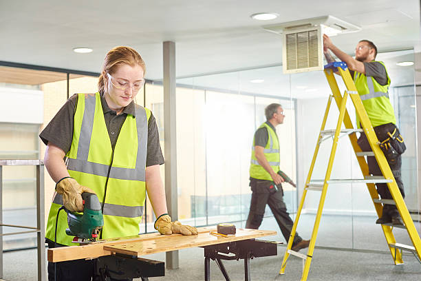female carpenter office fitout a teenage female carpenter is sawing studs on her workbench in a modern office refurbishment. IN the background a co-worker can be seen up a ladder working on the air conditioning unit. They are wearing safety workwear. train air conditioner stock pictures, royalty-free photos & images