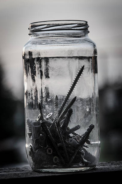 Defocus closeup shot through blurry thin glass jar with old metal bolts, nuts, washers and dowels. Grunge metal fastener fittings with blurred edges of frame