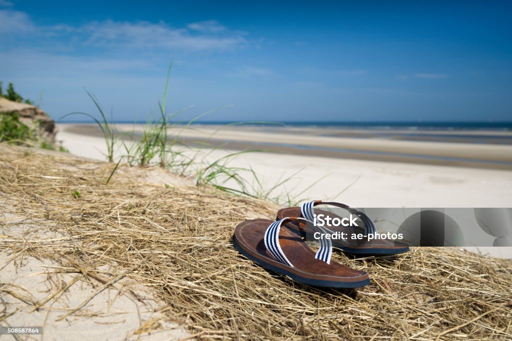 View of beach with flip-flops on sand dune View of Wangerooge beach under clear blue summer sky. Flip-flops and marram grass on a sand dune, shot with selective focus. Sandbar and north sea waves of the Wadden Sea UNESCO World Heritage Site in the background. Taken in Wangerooge Island, Friesland, Lower Saxony, German North Sea Region, Germany. Wangerooge Stock Photo