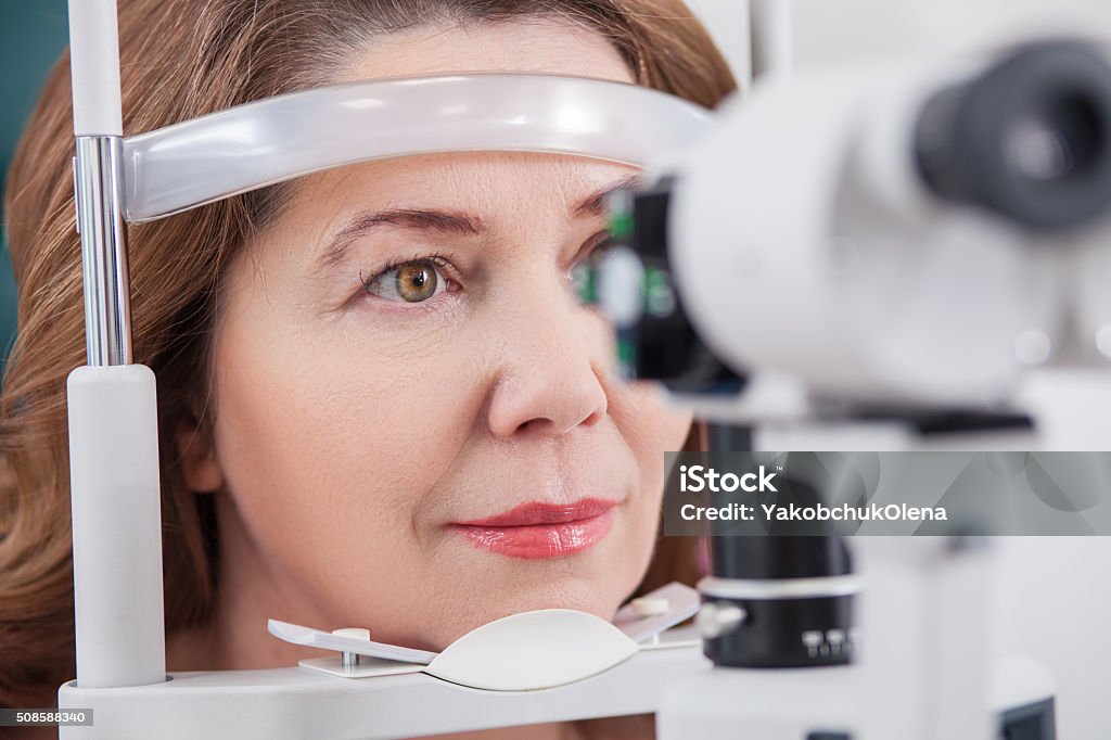 Cheerful lady having eye examination in oculist office Pretty woman is looking into eye test machine with concentration in oculist lab. Focus on her face Adult Stock Photo