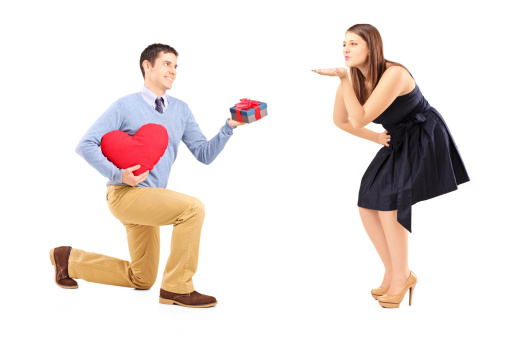 Smiling male on kneel with a red heart giving a present to a woman blowing him a kiss isolated on white background