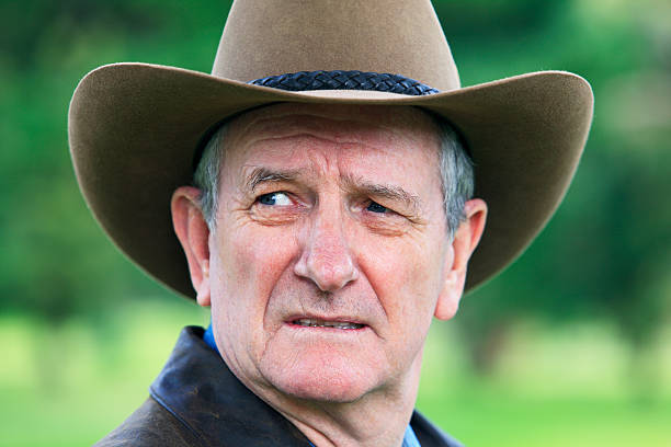 Head shot of a ruggedly handsome mature stockman (farmer, rancher) wearing a bush hat and appearing very worried while looking over his shoulder at something in the distance.  He is wearing waterproof coat with collar turned up for protection against the weather.  With blurred green bushland background.