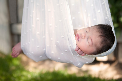 Newborn baby sleeping in white sling with rustic wooden fence in background.