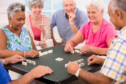 Multi-ethinc group of happy senior adult friends playing dominoes at local community center or friend's home.  Selective focus on dominos in center of table.