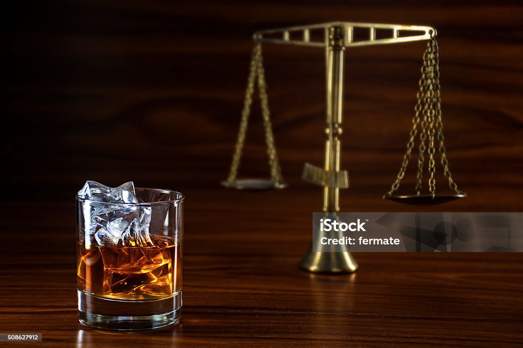 whiskey  and blurred scales of justice, concept alcohol and law whiskey glass with ice and the scales of justice blurred in the background on a wooden table, concept alcohol and conflicts with the law Alcohol - Drink Stock Photo