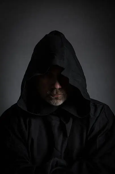 Head shot of a black man with hood hiding his face and showing only nose and chin with beard, in the shadow, on gray background, vertical