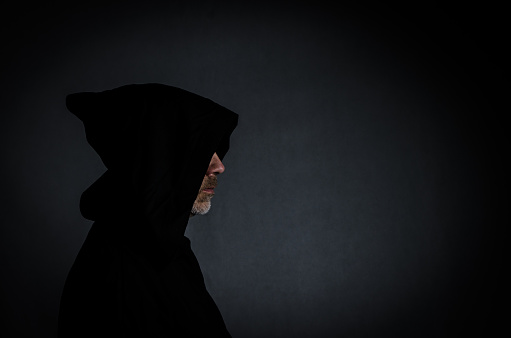Sideview head shot of a black man with hood hiding his face and showing only nose and chin with beard, in the shadow, on black background, horizontal.