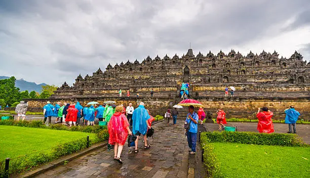 A group of tourist wearing colorful rain ponchos in front of Borobudur Temple. Borobudur is a 9th-century Mahayana Buddhist Temple in Magelang, Central Java, Indonesia. The monument consists of six square platforms topped by three circular platforms and is decorated with 2,672 relief panels and 504 Buddha statues. A main dome, located at the center of the top platform, is surrounded by 72 Buddha statues seated inside a perforated stupa. It is the world’s largest Buddhist temple, as well as one of the greatest Buddhist monuments in the world.
