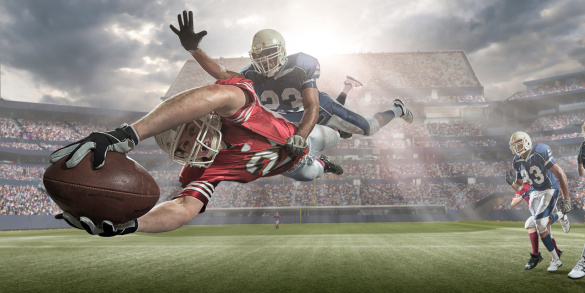 A close up image of an American football player in mid air about to score touchdown, whilst being tackled by rival player. Action takes place in a generic outdoor American football stadium under a cloudy sky with very bright and hazy evening sun. All players are wearing generic unbranded kit. 