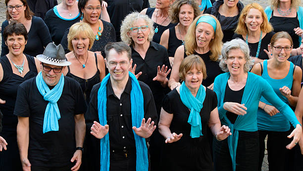 A group of people, singing men and women, dressed in black and blue or turquoise, standing, smiling,  performing, waving arms and hands, and looking at camera. It is a close-up of an almost white gospel choir.