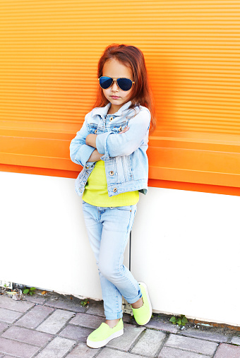Stylish little girl child wearing a sunglasses and jeans clothes over orange background
