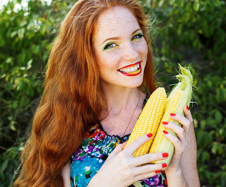 Redheaded sunny girl with many freckles on her face is holding an appetizing corn cob