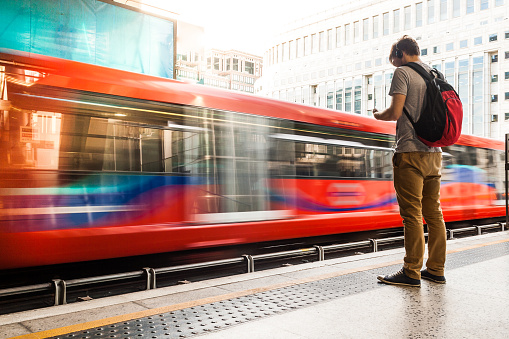 A photo of a young man wearing headphones while waiting for the train. He is on the right side of the image, and a red train is moving in front of him. The train has long windows that reflect the buildings in front of them. The middle section of the train is moving in a blur. There are truncated domes on the sidewalk between the man and the train. The man has short brown hair, and his headphones are black. He is wearing a gray short-sleeved shirt, tan-colored pants, black sneakers and a red and black backpack. A building with many windows is on the other side of the train. He is looking down at his smartphone. Horizontal colour image with copy space.