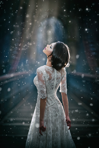 Lovely young lady wearing elegant white dress enjoying the beams of celestial light and snowflakes falling on her face. Pretty brunette girl in long wedding dress posing on a bridge in winter scenery