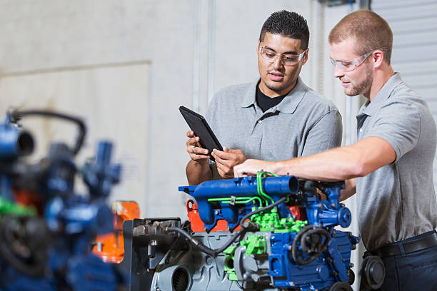 Two multi-racial men in auto mechanic school with engine Two multi-ethnic young men in vocational school, taking a class on reparing diesel engines.  They are working on an engine that has had parts painted different colors for training purposes.  They are wearing safety glasses. The Hispanic man is reading a digital tablet. multiengine stock pictures, royalty-free photos & images