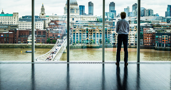 Rear view image of a businessman looking out at the modern London skyline from high up in his office building. He is looking across the river Thames, across the Millennium Bridge to St Paul's cathedral and the high rise skyscrapers of the City of London. He has his hands folden in front of him. Horizontal colour image with copy space.