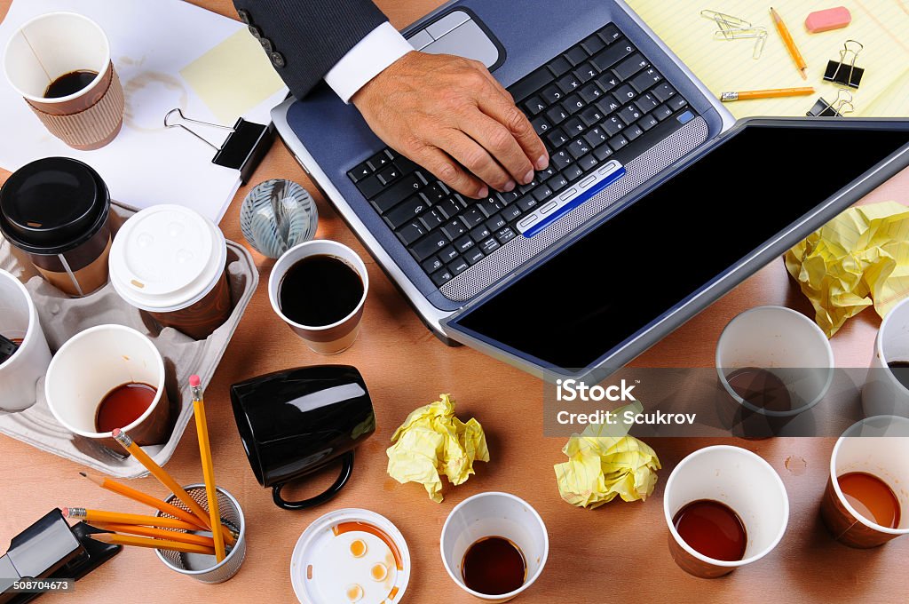 Cluttered Businessman's Desk Closeup view of a very cluttered businessman's desk. Overhead view with man's hand on laptop keyboard and scattered coffee cups and office supplies. Horizontal format. Stained Stock Photo