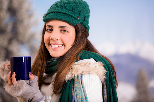 Latin descent young woman happily drinks a cup of hot coffee outside during the winter season.  Mountain scene in background.  She smiles as she looks at camera. Content, relaxed. Blue mug. Green knit cap, scarf. Mountains.