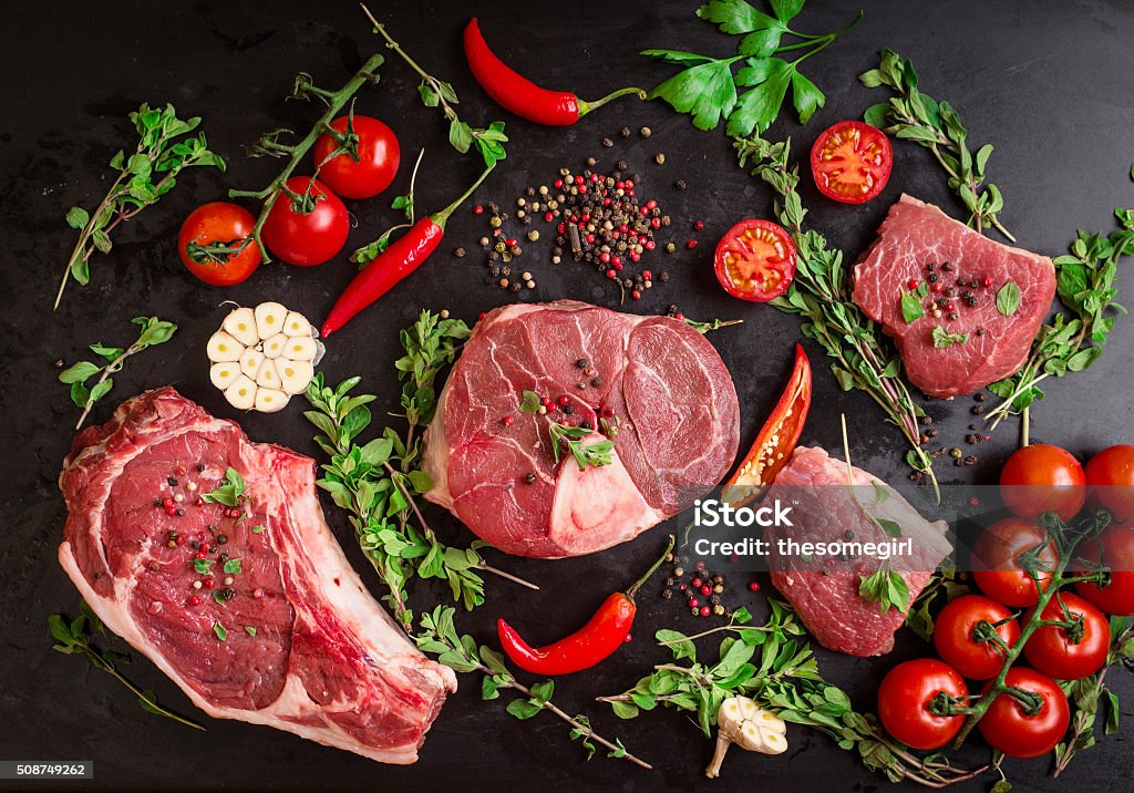 Raw meat steaks on a dark background ready to roasting Different types of steaks set. Assorted raw meat on a black chalk board background. Rib eye steak on the bone, veal shank (ossobuco), fillet with cherry tomatoes, hot pepper and herbs. Meat Stock Photo