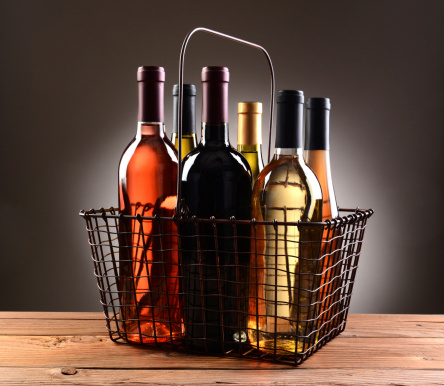 A wire shopping basket filled with assorted wine bottles. The basket is sitting on a rustic wooden table with a light to dark gray spot background.