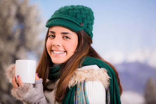 Latin descent young woman happily drinks a cup of hot coffee outside during the winter season.  Mountain scene in background.  She smiles as she looks at camera. Content, relaxed. White mug. Green knit cap, scarf. Mountains.