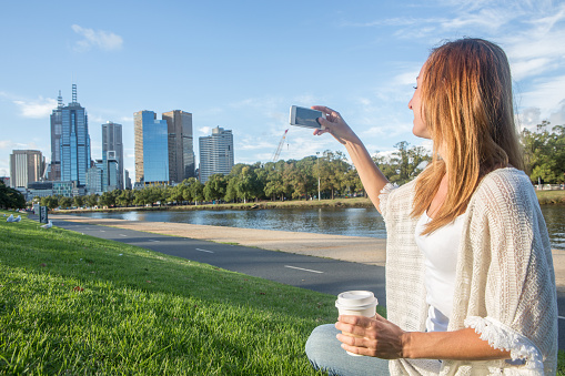 Cheerful young woman in Melbourne park seats on the grass, photographs the Melbourne CBD along the Yarra River on a beautiful day using a mobile phone. Australia.