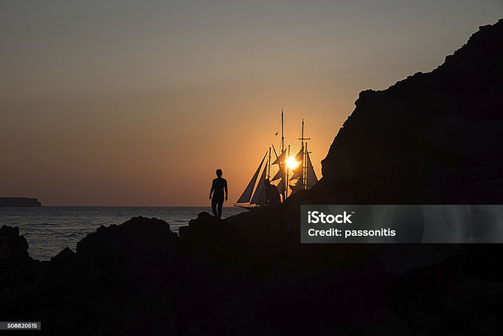 Silhouettes of a couple and sailing boat at sunset Passengers from the yacht disembarked on volcanic shore to watch the sunset Sailboat Stock Photo