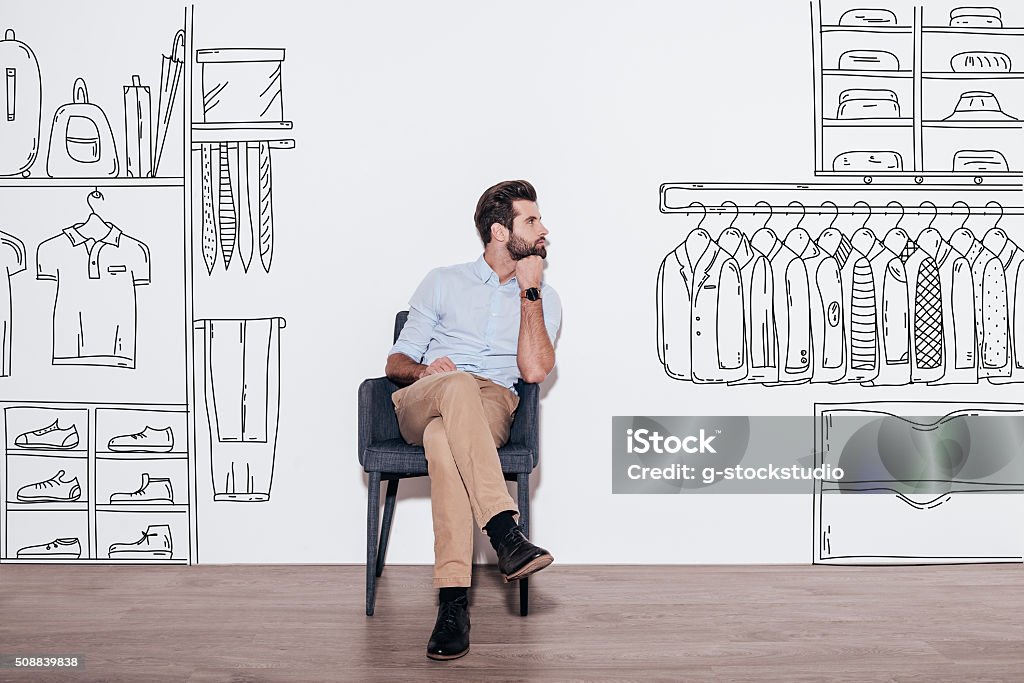 Dreaming about new wardrobe. Young handsome man keeping hand on chin and looking away while sitting in the chair against illustration of closet in the background Closet Stock Photo