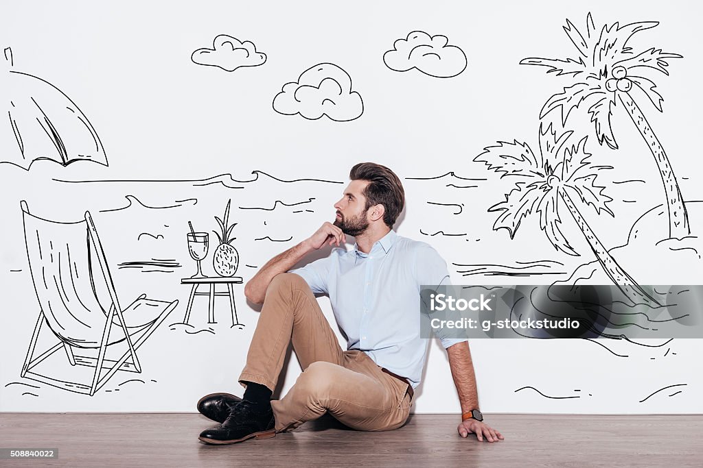 Dreaming about vacation. Young handsome man keeping hand on chin and looking away while sitting on the floor with illustration of resort in the background Dreamlike Stock Photo