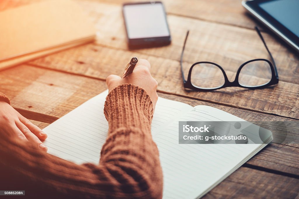 Planning new day. Close-up image of woman writing in notebook with copy space while sitting at the rough wooden table Writing - Activity Stock Photo