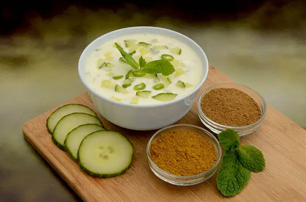 A bowl of freshly made spicy cucumber raita surrounded by its ingredients of cucumber, mint, coriander and cumin.