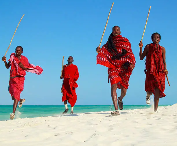 Four Maasai warriors running on the beach with their traditional sticks held in the air. They are all dressed in traditional maasai red robes. They are running on the white sand on shores of Indian ocean.