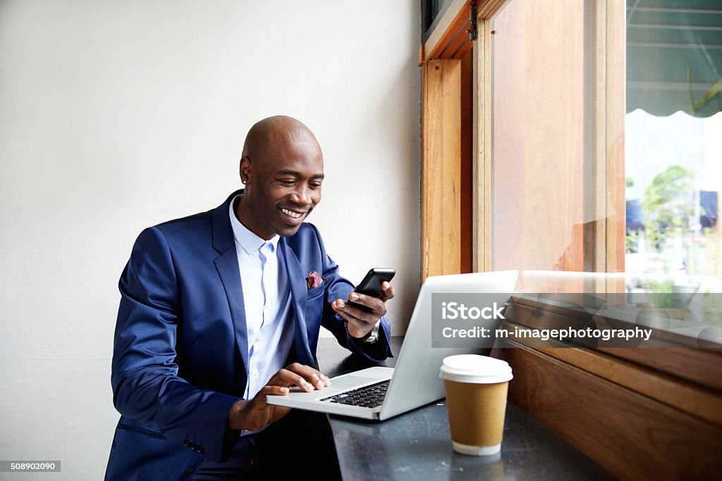 Happy african businessman using phone at cafe Portrait of happy african businessman using phone while working on laptop in a restaurant On The Move Stock Photo