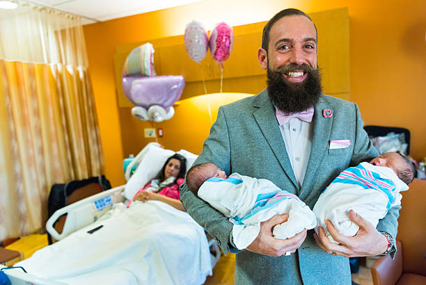 Proud father of twins Smiling bearded caucasian man posing with his twin newborn babies with his wife in the background at the hospital room unknown gender stock pictures, royalty-free photos & images