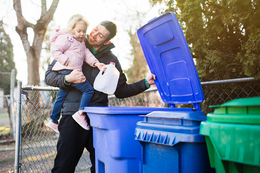 A dad holds his daughter as he teaches her about the environmentally friendly practice of recycling plastic and cardboard waste.  A good practical learning opportunity.