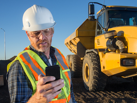 A royalty free image of a construction engineer at a construction site in front of a hauling truck using a smart phone to text, make a call, use an app, check email or communicate. The blue or white collar worker is wearing a hardhat, safety glasses and safety vest.