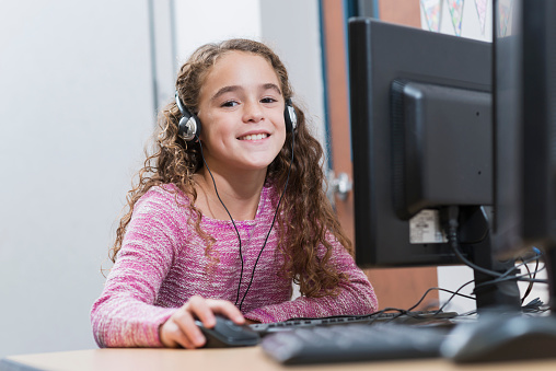 A mixed race Hispanic 9 year old girl using a desktop computer at school. She is wearing headphones, using the mouse, looking at the camera and smiling.