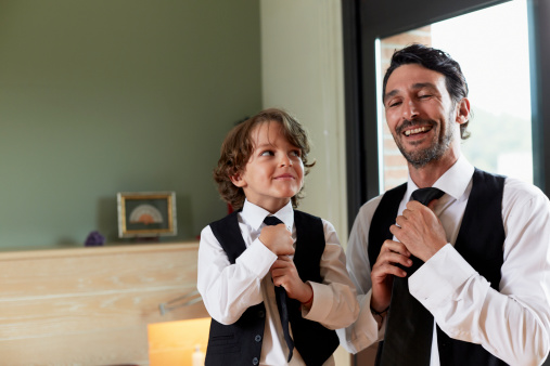 Cute little boy adjusting tie while looking at father in house