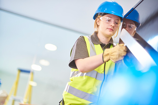 a teenage female apprentice carpenter is installing handrails  in a modern office refurbishment. Her colleague is in the background holding building plans . She is using a drill to make guide holes for the hand rails  . They are wearing safety workwear.