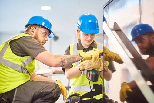 a teenage female apprentice carpenter is installing handrails  in a modern office refurbishment. She is being supervised by her co-worker . She is using a drill to make guide holes for the hand rails  . They are wearing safety workwear.
