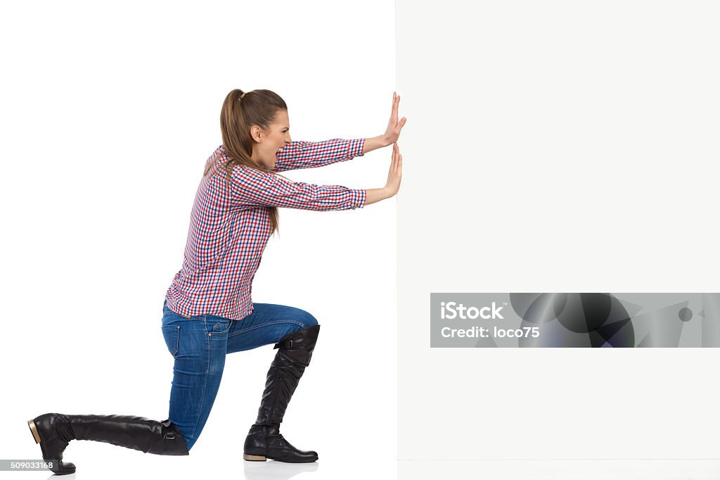 Woman In Lumberjack Shirt Pushing Wall Young woman in jeans, black boots and lumberjack shirt pushing a white wall and shouting. Side view, full length studio shot isolated on white. Pushing Stock Photo