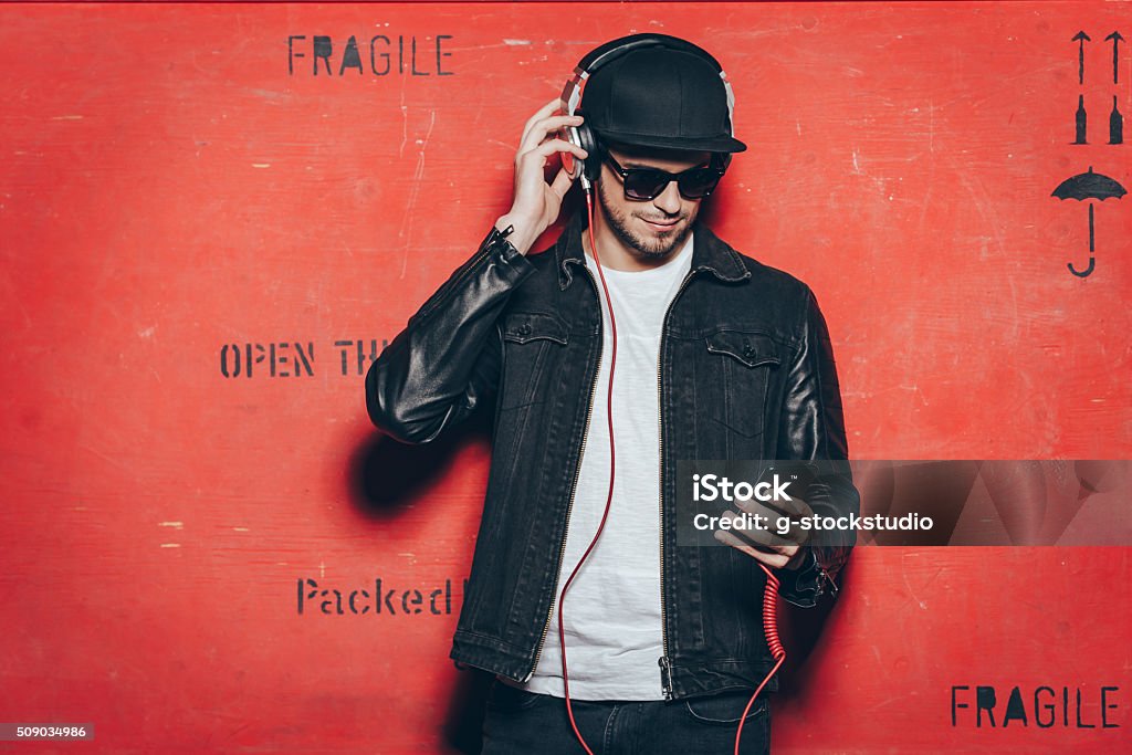 Style and music. Handsome young man adjusting his headphones and looking at his smart phone while standing against red background Men Stock Photo