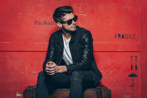 Handsome young man in sunglasses keeping hands clasped and looking away while sitting on wooden chest against red background