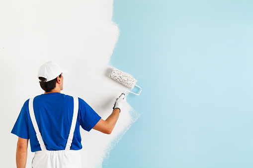 Back view of  painter in white dungarees, blue t-shirt, cap and gloves painting a wall with paint roller, with copy space